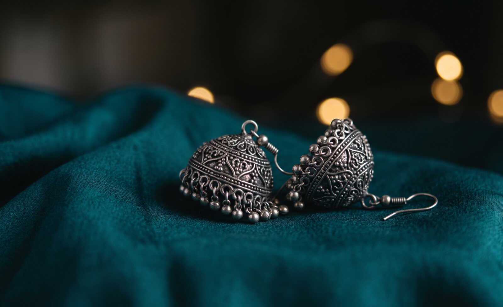Oxidised silver jhumka earrings resting on teal silk, warm fairy lights glowing behind