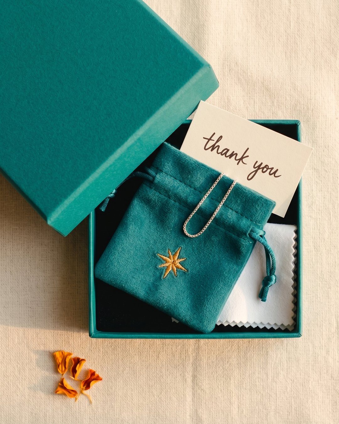 Jonaki packaging — a teal velvet pouch with embroidered amber spark inside a teal gift box, with a handwritten thank-you card on cream linen